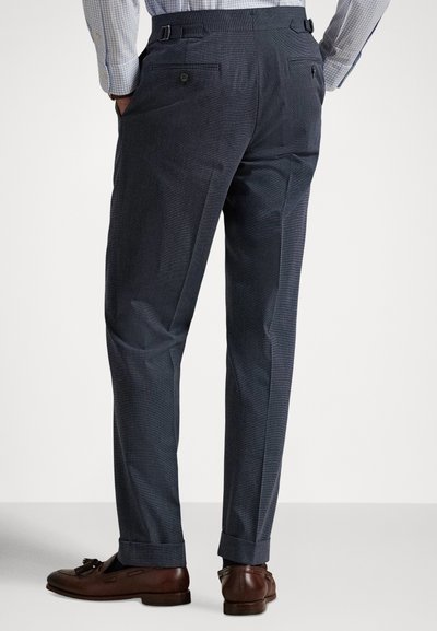 Vyras dėvi pritaikytas tamsiai mėlynas kelnes su atvartais, rudos odos laisvus batus su kuodeliais ir šviesiai mėlyną languotą marškinių su kišenių įdėjimu į kelnes, rankos kišenėse.