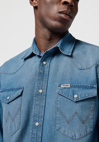 Camisa de mezclilla azul claro con cuello puntiagudo, dos bolsillos en el pecho, botones a presión y costuras en marrón que contrastan. Logo de Wrangler en el bolsillo.