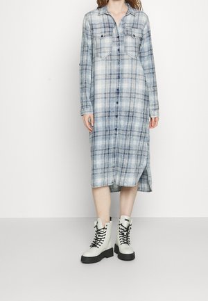 Blauwe geruite blousejurk met twee borstzakken, zijsplitten en lange mouwen, gecombineerd met witte veterschoenen.