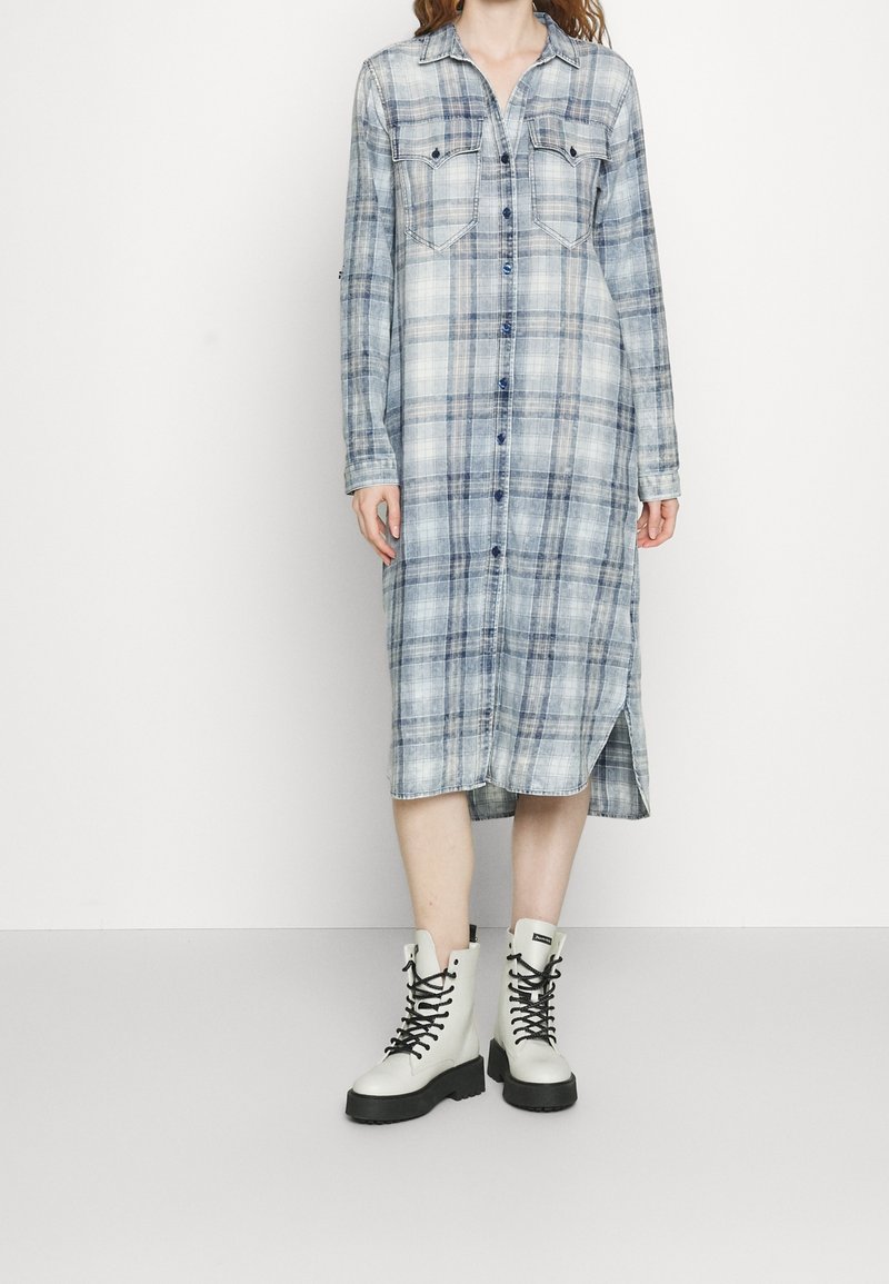Niebieska koszulowa sukienka w kratę z dwiema kieszeniami na piersi, rozcięciami po bokach i długimi rękawami, połączona z białymi lace-up butami wojskowymi.