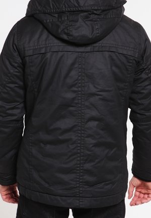 Persoon in een zwart hoodie-jack, van achteren weergegeven met ontspannen handen langs de zij tegen een effen achtergrond.