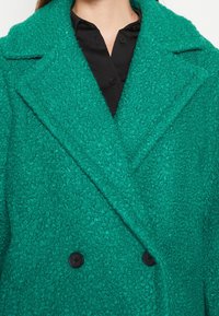 Femme portant un manteau double boutonnage texturé bleu sarcelle sur une chemise noire boutonnée, coupée au niveau du cou et du milieu du torse.