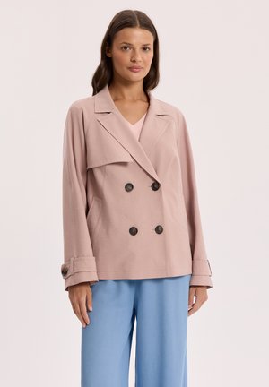 Kobieta ubrana w bladoróżową marynarkę dwurzędową z mankietami zapinanymi na guziki oraz jasnoniebieskie szerokie spodnie na tle gładkiego tła.