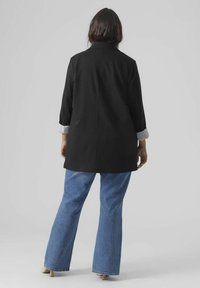 Blazer noir à manches trois-quarts, avec des poignets rayés. Associé à un jean évasé bleu clair. Fond neutre.