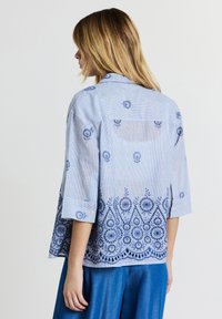 Frau in einer hellblauen Bluse mit blauer floraler Stickerei und hochgerollten Ärmeln, kombiniert mit einer lockeren blauen Hose, seitlich blickend.