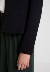 Blazer noir texturé avec un ourlet arrondi et une coupe légèrement ample, associé à un pantalon vert taille haute avec des boutons dorés.