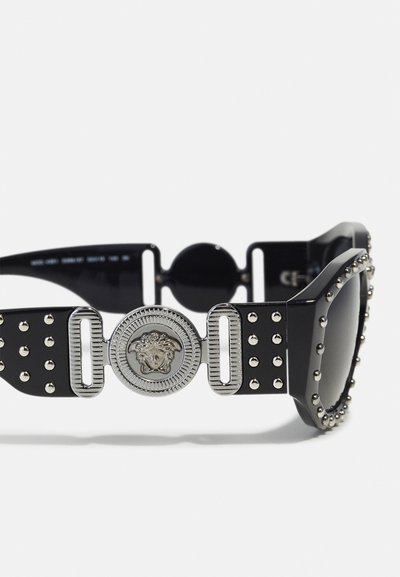 Black sunglasses with a thick frame, adorned with silver studs and a prominent silver lion emblem at the temple. Square shape, dark lenses.