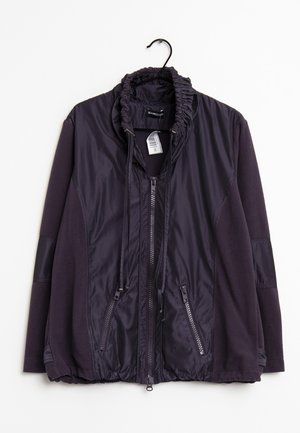 Veste zippée violet foncé avec col élastique froncé, poches zippées et manches longues, suspendue à un cintre noir sur fond blanc.