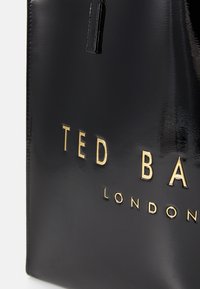 Tote en cuir verni noir avec une texture lisse, arborant une inscription en lettres dorées et une fine poignée. Design simple, sans motifs visibles.