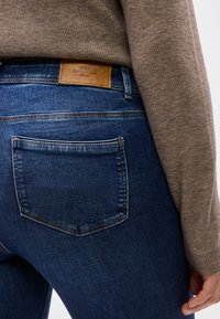 Κοντινό πλάνο ενός ατόμου που φοράει σκούρο μπλε τζιν τζιν με πίσω τσέπη και ταμπά δερμάτινη ετικέτα μάρκας, συνδυασμένο με καφέ πλεκτό πουλόβερ.