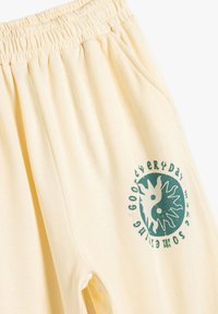 Pantalones de chándal amarillos claros con una cinturilla elástica. Presenta un gráfico circular verde con diseños florales en el muslo izquierdo. Tejido suave.