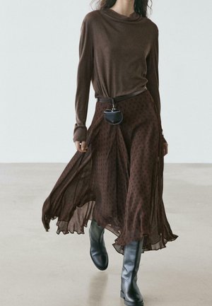 Femme portant une jupe fluide marron foncé à motifs, un haut marron à manches longues, une petite pochette noire à ceinture et des bottes noires montant jusqu'aux genoux, marchant à l'intérieur.