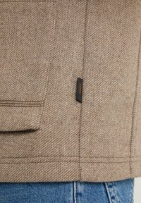 Brun herringbone-mönstrad jacka med en texturerad yta och ficka med klaff, med en svart etikett med orange text på sömmen.