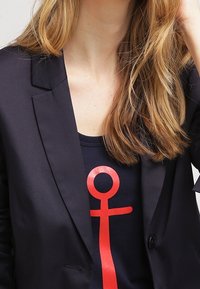 Svart skräddarsydd blazer över en svart skjorta med en stor röd ankh-symbol. Slät tyg med en strukturerad passform och en knappdetalj.