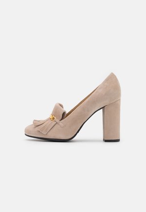 Zapato de tacón alto de ante beige con punta redondeada, detalle de flecos y un acento de hardware en tono dorado. Presenta un tacón grueso para mayor estabilidad.