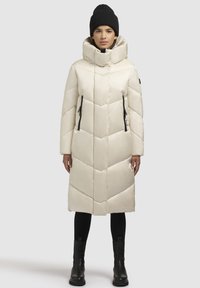 Cremefarvet puffjakke med høj krave, chevron-mønster, lynlåslommer og lang silhuet, båret med sorte støvler og leggings.