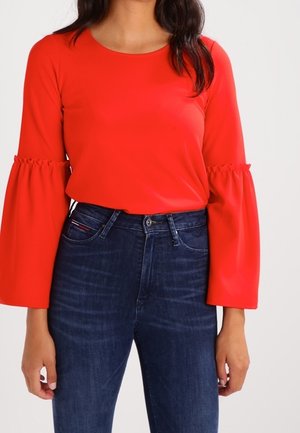 Kvinde iført en skrigrød bluse med opsamlede klokkeærmer og mørkeblå højtaljede jeans, stående mod en ensfarvet baggrund.