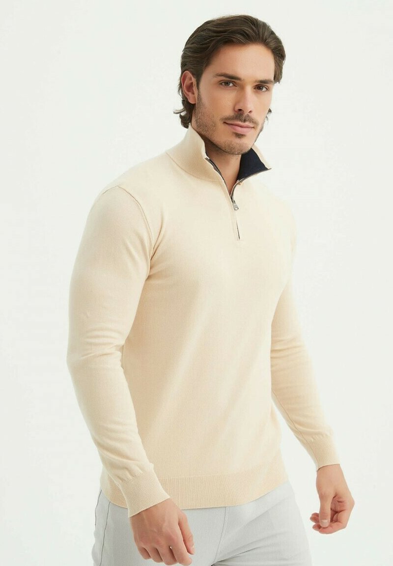 Pull en tricot beige avec un col haut et une fermeture éclair. Manches longues et poignets côtelés, présentant un intérieur contrastant en bleu marine au niveau du col.
