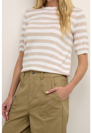 Vrouw draagt een beige-wit gestreept T-shirt met korte mouwen en een hoge taille khaki broek, met één hand in de zak, tegen een effen achtergrond.