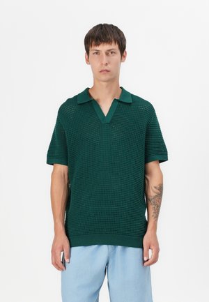 Green knitted polo shirt with a textured pattern, short sleeves, and a collared neckline, paired with light blue trousers.