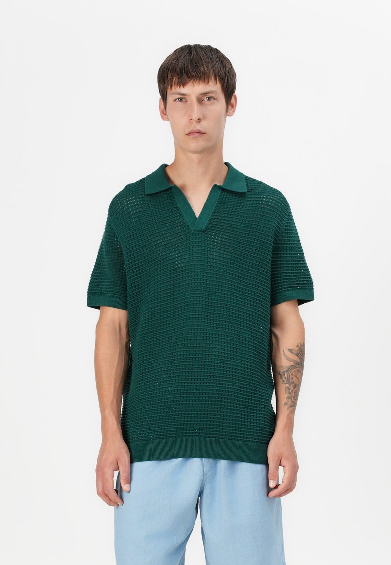 Polo shirt tricoté vert avec un motif texturé, à manches courtes et col, associé à un pantalon bleu clair.