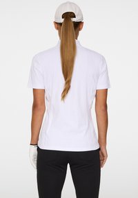 Korte mouwen witte polo met een getextureerd golfpatroon, achteraanzicht. Gecombineerd met zwarte broek, accessorized met een handschoen en een witte pet.