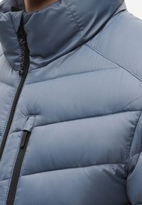 Közeli kép egy világoskék steppelt dzsekiről, magas gallérral és fekete cipzárral a mellzsebnél és az elülső részen.