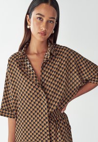 Bruine bloezen met zwart geometrisch patroon, korte mouwen en een knoopsluiting aan de voorkant. Beschikt over een elastische taille voor aanpassing van de pasvorm.