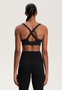 Črn športni nedrček s križnimi naramnicami in nastavljivimi kljucki na zadnji strani; gladka tkanina, minimalno brandiranje, v kombinaciji s črnimi visokim pasom leggingi.