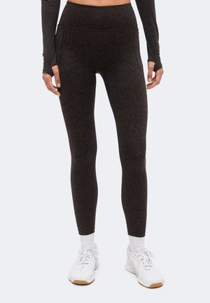 Vrouw draagt hooggetailleerde donkere legging met slangprint, witte enkelsokken, witte sportschoenen en een zwart crop top met lange mouwen, staand.