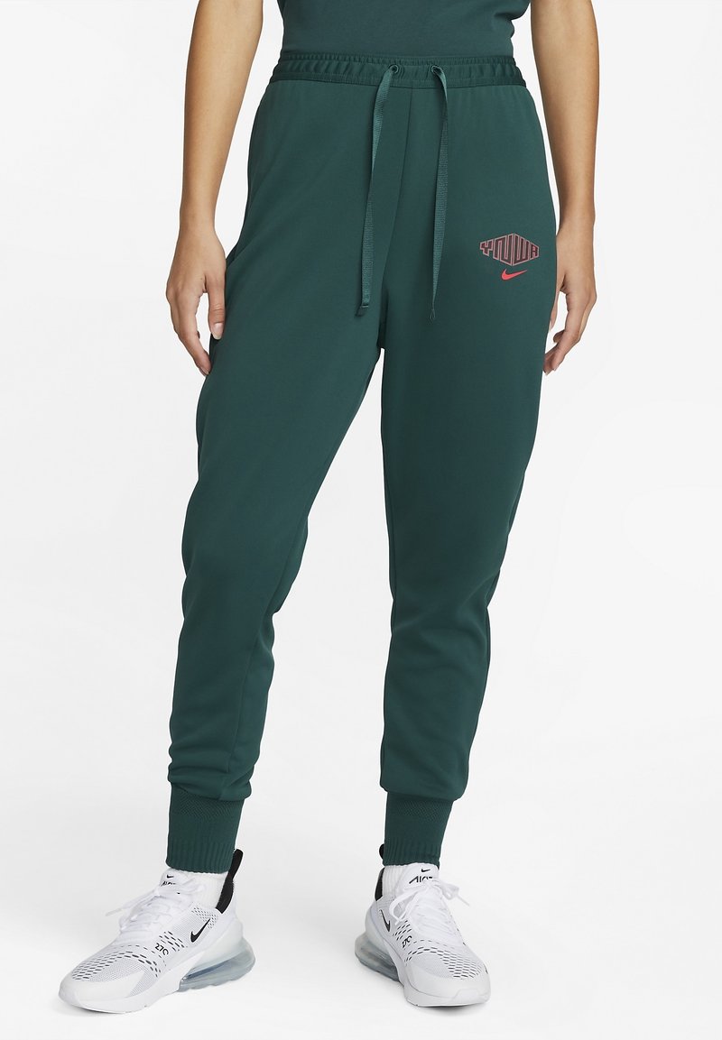 Joggers vert foncé avec taille élastique et cordon de serrage, logos rouges "Puma" et Nike sur la cuisse, portés avec des baskets blanches Nike Air.