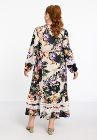 Langes Blumenmaxikleid mit mehrfarbigen Blumen auf beigem Hintergrund, mit langen Ärmeln und Spitzenakzenten am Saum. Sandalen mit Blockabsatz.