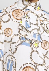 White blouse featuring a rope and compass print, with blue and yellow accents and a tie collar. Buttons down the front.