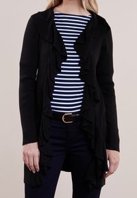 Svart cardigan med volangkant på framsidan, stickad textur, bärs över en marinblå och vit randig skjorta. Har ett bälte med en guldfärgad spänne.