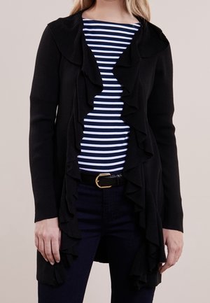Sort cardigan med rynkede forkanter, strikket tekstur, båret over en marineblå og hvid stribet skjorte. Har et bælte med en guldspænde.