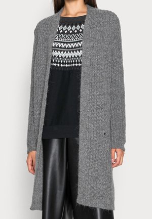 Femme portant un long cardigan gris tricoté, un pull noir à motifs et un pantalon en cuir noir, debout devant un fond uni.