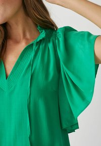 Zelená blúzka s výstrihom do V, na pleciach zdobená nariasením a s krátkymi volánovými rukávmi, vyrobená z ľahkej, hladkej látky.