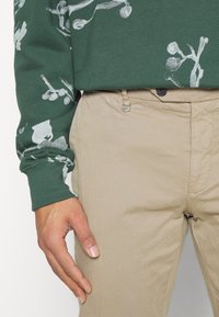 Chemise verte à manches longues avec impression florale blanche, associée à un pantalon beige avec une poche avant et un détail métallique.
