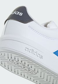 Basket blanche Adidas avec bande bleue, languette de talon noire avec logo Adidas, et semelle texturée avec marquage en relief Adidas.