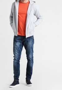 Felpa zip grigia chiara, t-shirt arancione, jeans blu, sneakers blu scuro. Tessuto liscio, design informale, vestibilità standard.