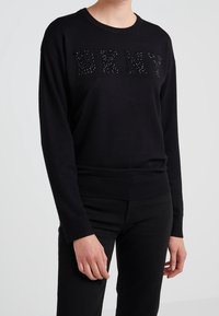 Svart stickad tröja med bokstäverna "DKNY" i strukturerad, pärlbelagd detalj. Ribbad mudd och fåll. Kombinerad med svarta byxor.