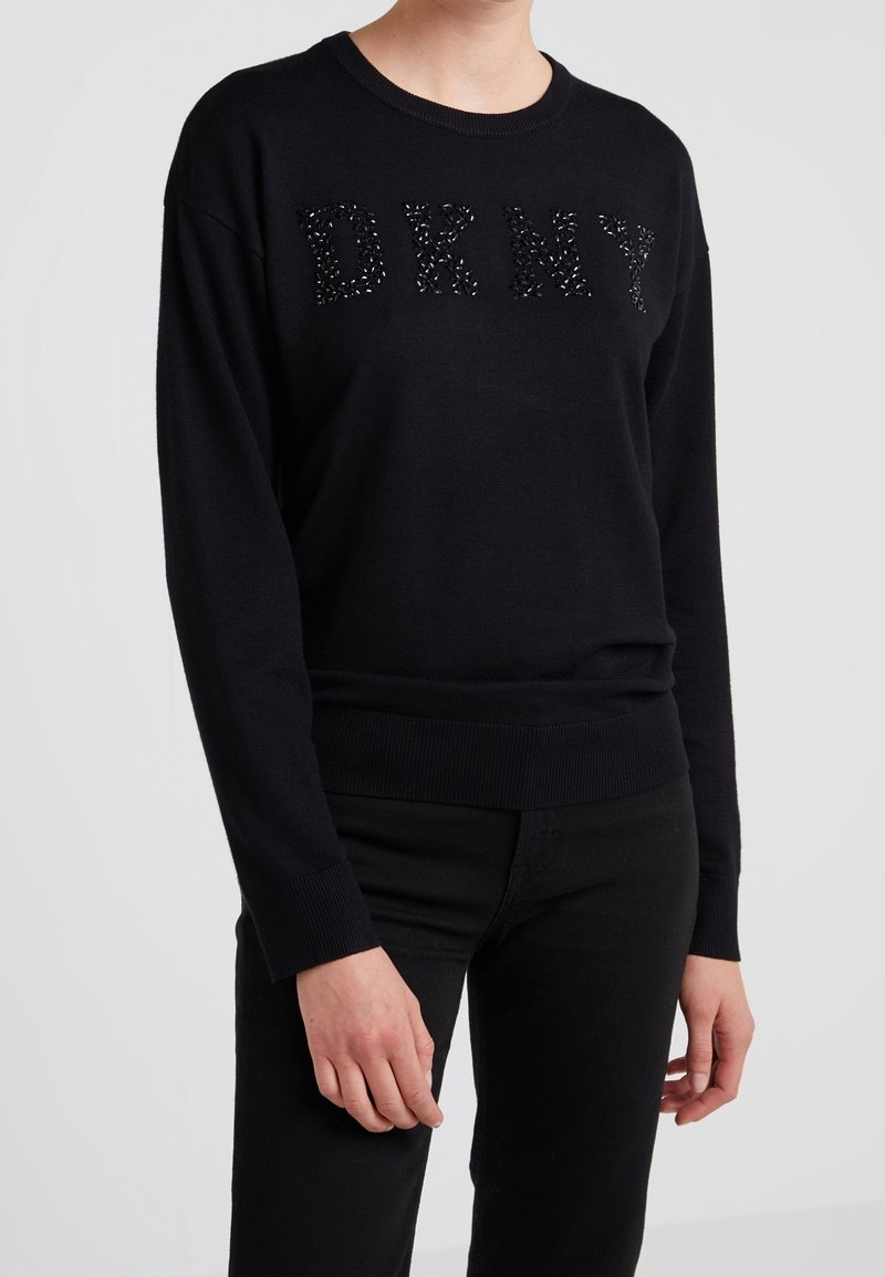 Svart stickad tröja med bokstäverna "DKNY" i strukturerad, pärlbelagd detalj. Ribbad mudd och fåll. Kombinerad med svarta byxor.