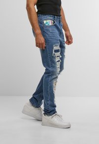 Bărbat îmbrăcat cu blugi bleumarin rupți, cu petice de material colorat, adidași albi și un tricou fără mâneci negru, stând în fața unui fundal simplu.