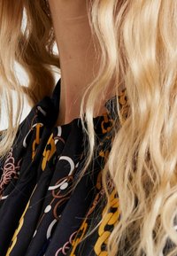 Blouse noire avec un motif chaîne en jaune, brun et blanc. Tissu texturé avec détail de col à volants et fermeture par boutons.