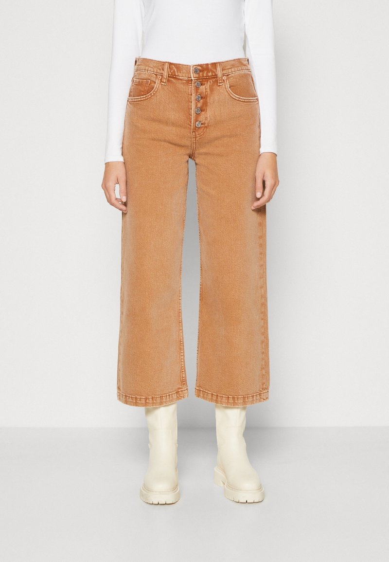 Pantalones de mezclilla de pierna ancha color marrón claro con cierre de botones, que cuentan con cinco bolsillos y un dobladillo sin terminar, combinados con botas de tobillo color crema.