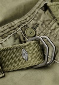 Olijfgroene canvas riem met een gestructureerd oppervlak, voorzien van een metalen gesp en een logo-label. Zichtbare taillebandstiksels op de stof.