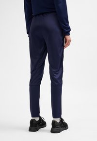 Granatowe spodnie sportowe o zwężanym kroju, charakteryzujące się gładką teksturą i bocznymi akcentami. Noszone z czarnymi butami do biegania.