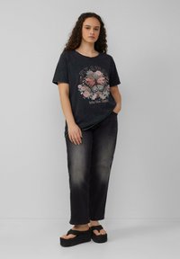 Zwart graphic T-shirt met een vlinder en bloemdessin, gedragen met donkere spijkerbroek en zwarte platform sandalen. Krullend haar.