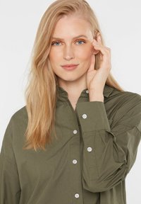 Olive green button-up shirt with a high collar, long sleeves, and white buttons; smooth fabric with a slightly glossy texture.