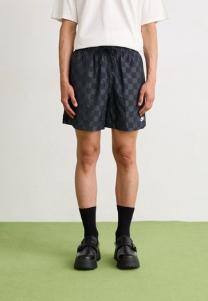 Zwart geruite zwembroek met een elastische tailleband, met een klein wit logo. Gecombineerd met zwarte sokken en stevige zwarte schoenen.